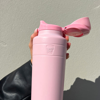 Hand with pink nail polish holding a Pashmak Pink Ora Bottle 700ml against a light grey textured wall, with the flip-top lid open to reveal the drinking spout, showing the embossed KeepCup logo on the insulated stainless steel body.