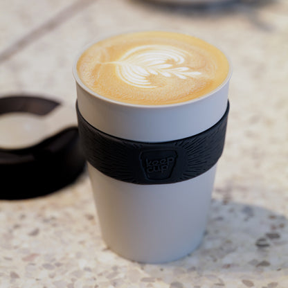White KeepCup reusable coffee cup with black silicone band containing latte with rosetta latte art on marble surface
