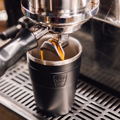 Espresso being poured into a black KeepCup stainless steel cup from a coffee machine.