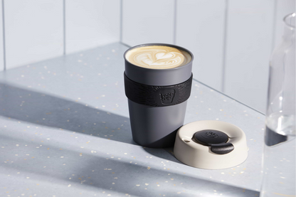 Grey KeepCup reusable coffee cup with black grip band containing latte with heart latte art, lid beside cup on speckled counter