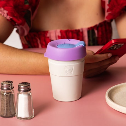 White KeepCup reusable stainless steel coffee cup with purple lid on a pink table, person using phone in background
